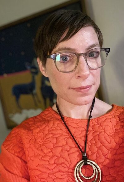Woman in glasses, orange shirt, and necklace in front a painting.