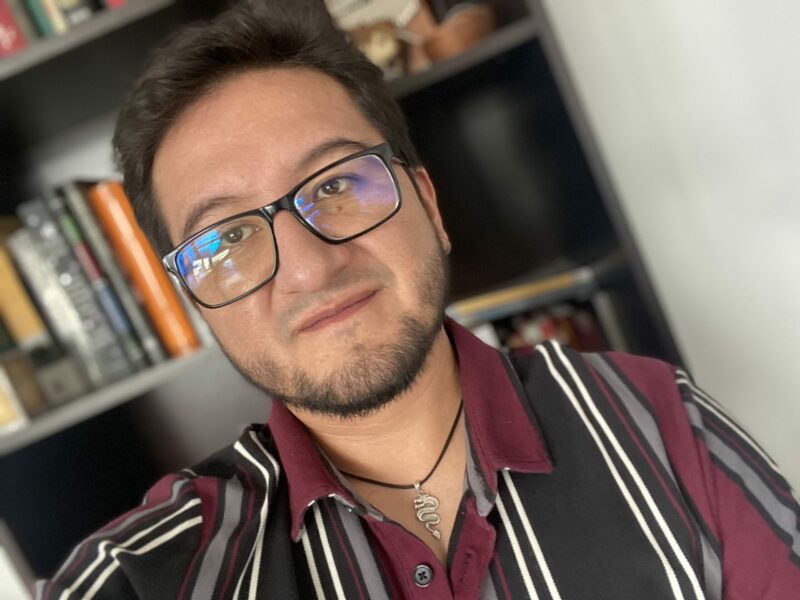 Daniel Romero-Alvarez Headshot, man in striped shirt with glasses in front of a bookcase filled with books.