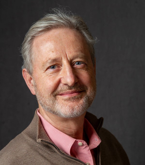 Man, Patrick Webb, in front of neutral dark gray background with pink collared shirt and a coat.